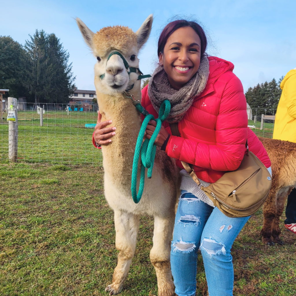 A woman in a red jacket smiling while hugging Sassafras, a cream-colored alpaca, during a farm experience from the "Adopt an animal" package.