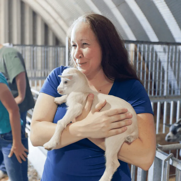 A smiling woman wearing a blue shirt cradles a baby goat in her arms inside a clean, spacious barn with gated enclosures in the background.