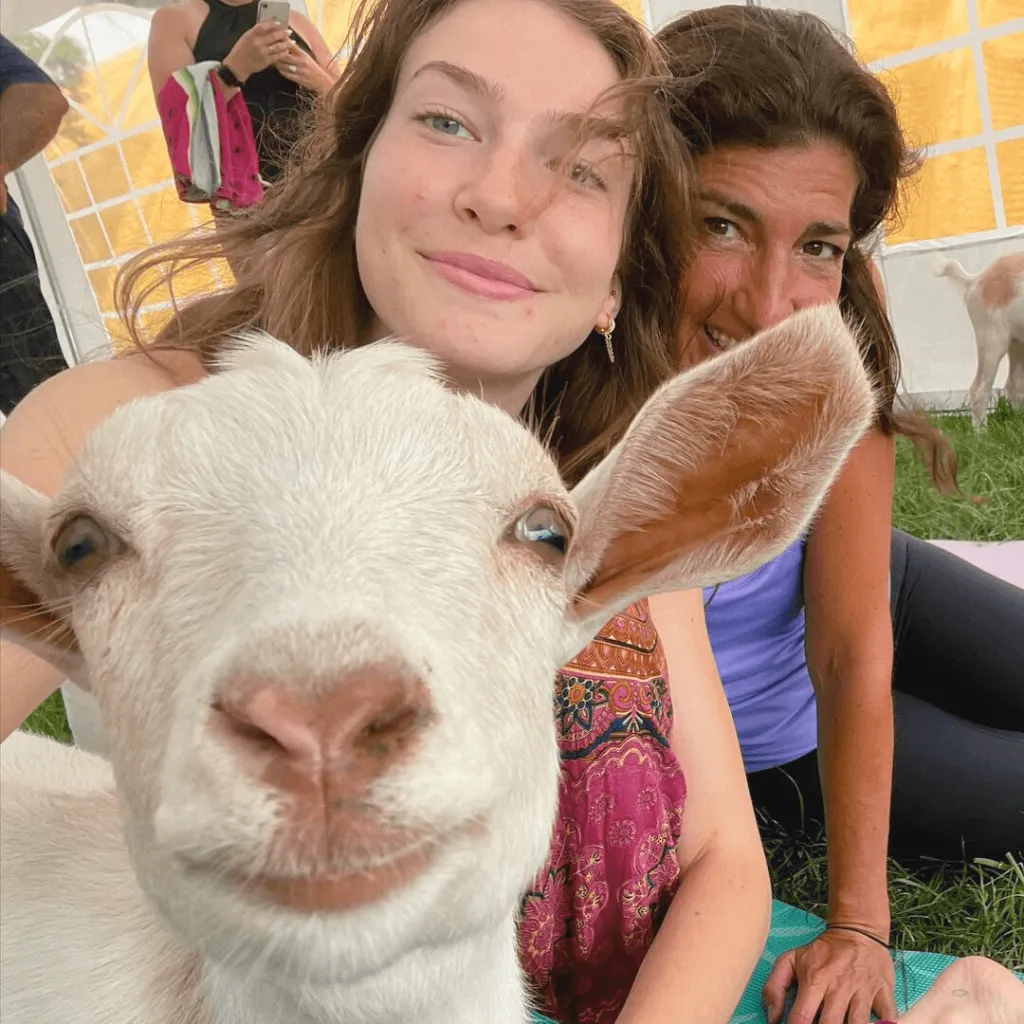 A close-up of a smiling goat with two participants enjoying goat yoga at Udderly Ridiculous Farm Life in the background.