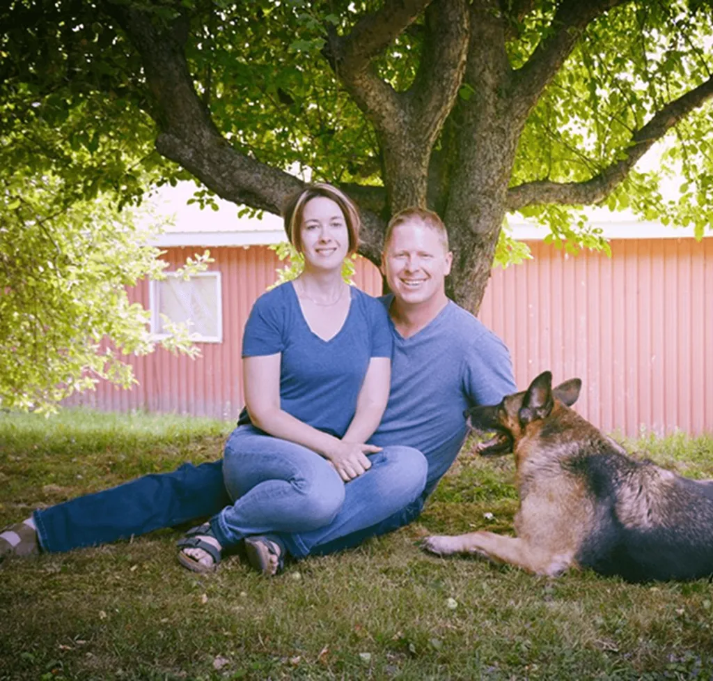Cheryl and Greg sitting together under a tree on their farm, smiling warmly with their loyal German Shepherd by their side. A peaceful moment reflecting the heart of Our Story.