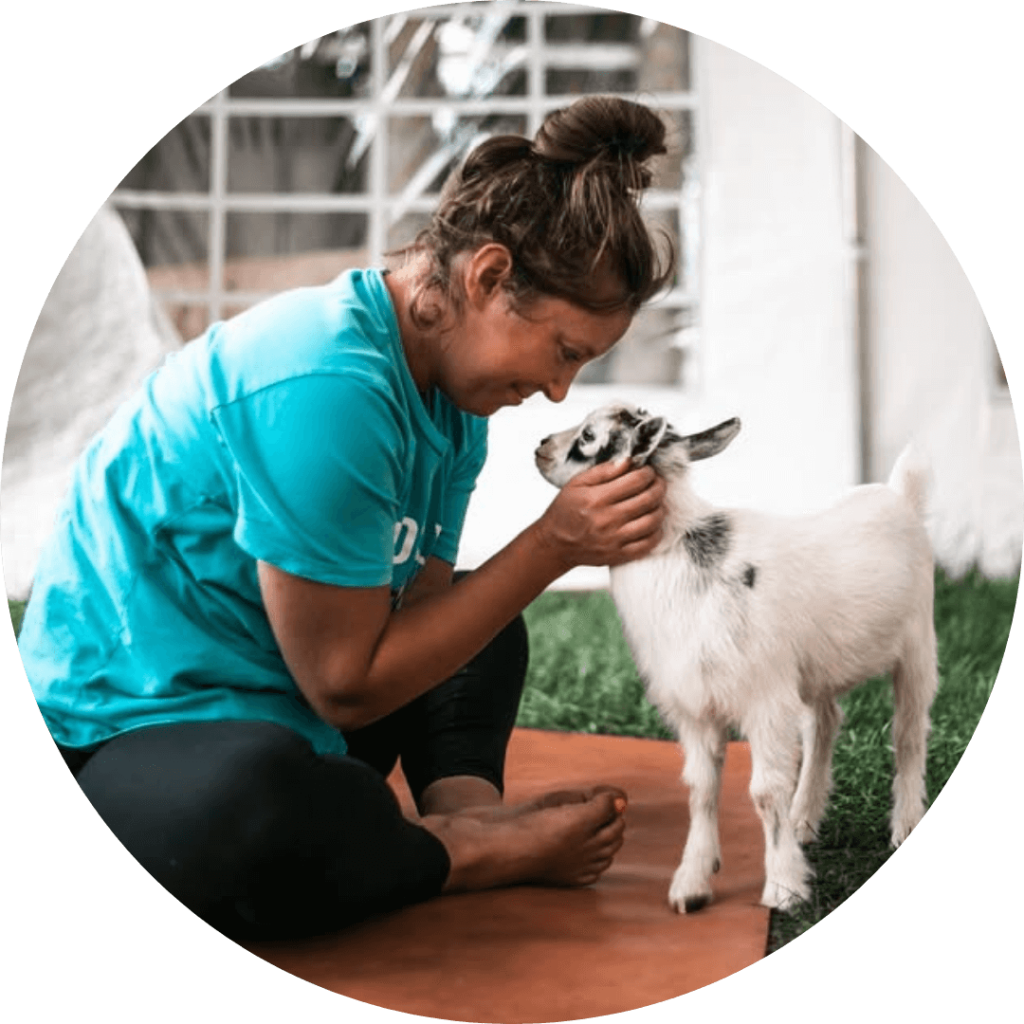 Cassie Scoyne smiling at a small goat while seated on a yoga mat, goat yoga instructor at Udderly Ridiculous.