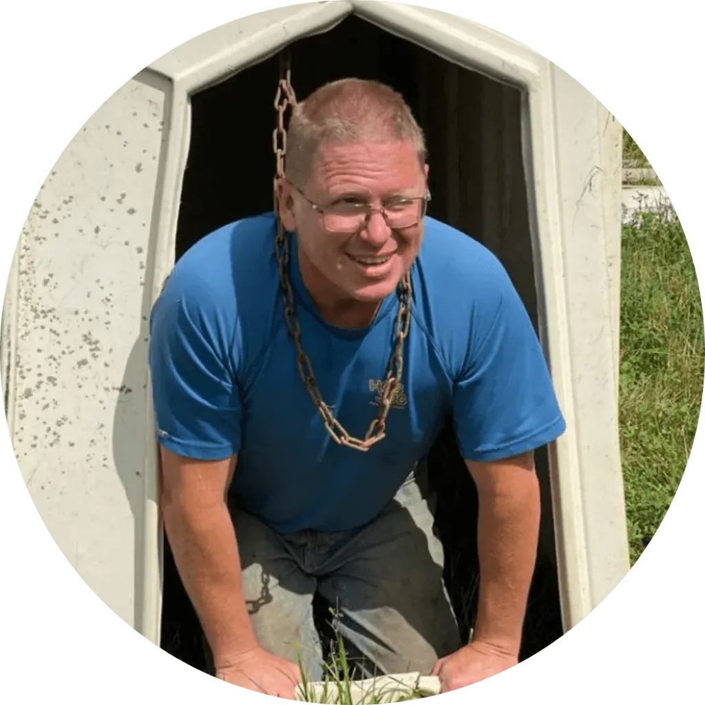 Greg Haskett smiling from inside a calf hutch, wearing a blue shirt and jeans, husband of Cheryl Hackett.